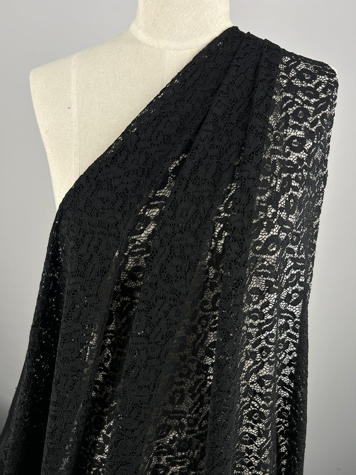 A lightweight Black Lace - Ora - 150cm fabric by Super Cheap Fabrics is draped over a dress form in a one-shoulder style, showcasing the intricate floral pattern. The sheer stretch material is semi-transparent, allowing light to pass through and highlight the detailed design. The background is a plain gray.