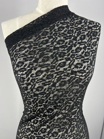 A mannequin is dressed in a black, one-shoulder lace dress with an intricate floral pattern made from Black Lace - Ora - 150cm by Super Cheap Fabrics. The lace overlays a nude-colored lightweight fabric, creating a contrast that highlights the details of the black lace. The background is neutral gray.