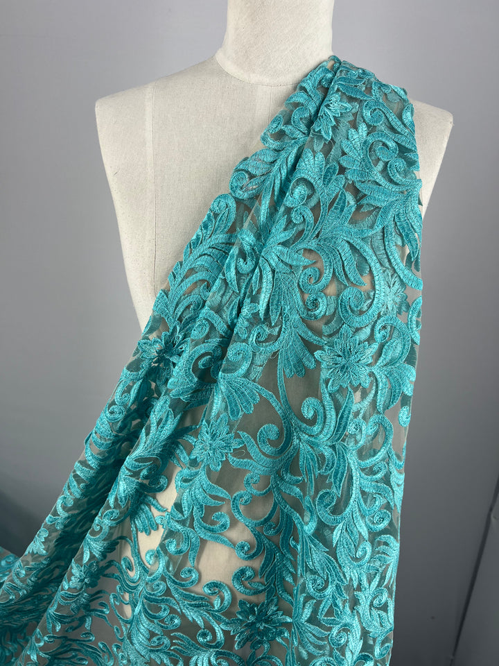 A mannequin draped in Super Cheap Fabrics' Evening Lace - Aqua Galadriel - 125cm with intricate, swirling floral patterns. The flower design's rich texture and vibrant color are prominently showcased against the neutral background.