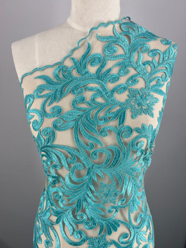 A dress form draped with Evening Lace - Aqua Galadriel - 125cm by Super Cheap Fabrics, featuring floral and swirling vine patterns. The fabric, accented with intricate applique, covers one shoulder and extends diagonally across the bust, revealing transparent mesh sections. The background is a plain gray.