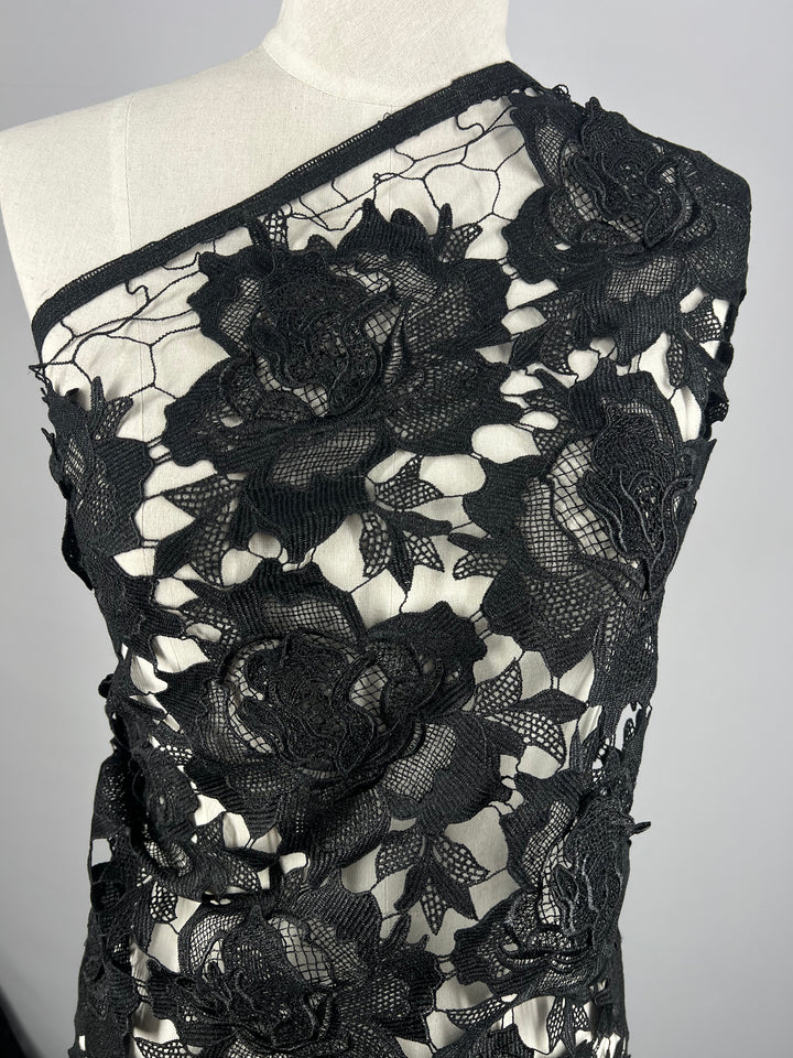 A close-up image of a mannequin draped in Designer Anglaise Lace - Black - 120cm by Super Cheap Fabrics featuring intricate floral patterns. Made from 100% polyester, the fabric's detailed roses and foliage create an elegant, textured design on a white background. The black fabric has a 120cm width, perfect for various crafting needs.