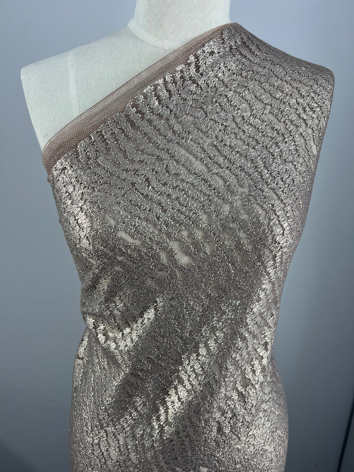 A dress form is draped in a luxurious, sheer metallic fabric that features intricate, shimmering patterns in a soft, champagne hue. The Evening Lace - Avalanche - 150cm by Super Cheap Fabrics is wrapped diagonally across the form, displaying its texture and elegant, reflective quality under the lighting.