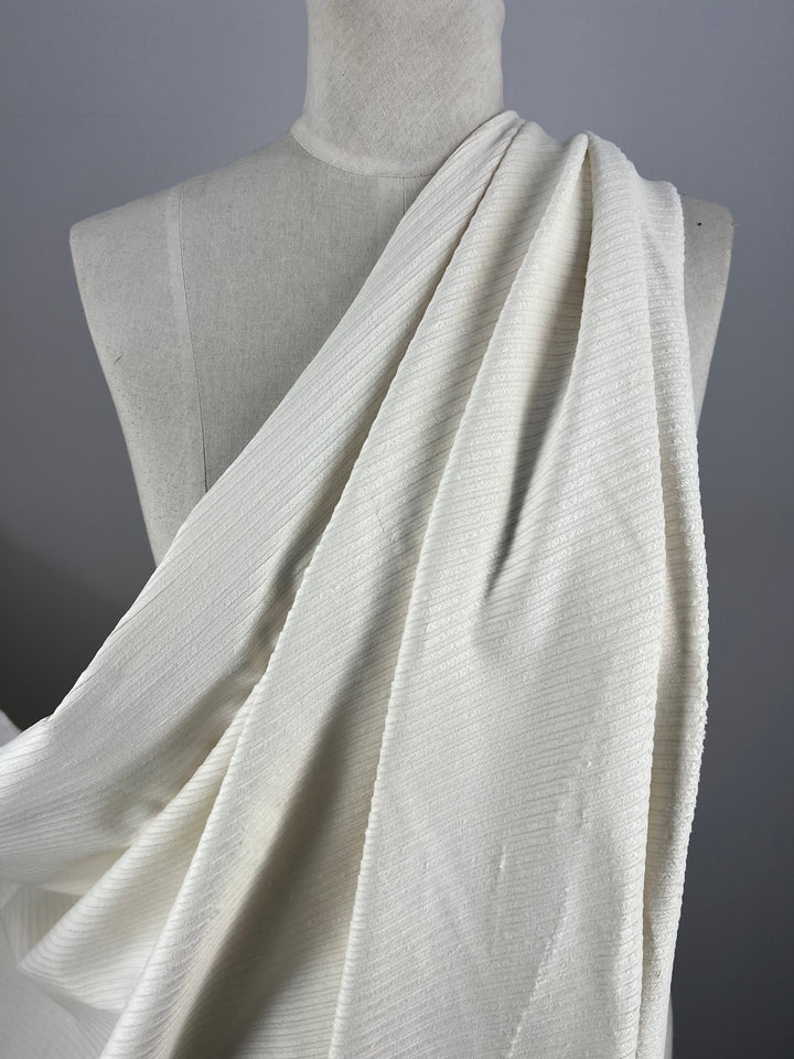 A close-up of a white dress form draped with Super Cheap Fabrics' Medium Wale Stretch Corduroy in Ivory. The textured material features subtle vertical lines, creating gentle folds over the form. This medium to heavy weight fabric stands out against a plain, light grey background, adding elegance and depth.