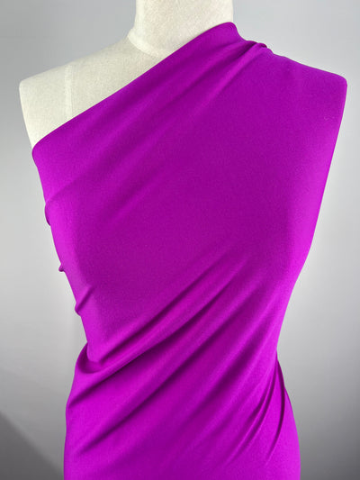A mannequin is dressed in a vibrant purple one-shoulder garment crafted from Super Cheap Fabrics' Plain Lycra - Lipstick, 155cm. The form-fitting garment is smooth with minimal wrinkles, set against a plain, light grey background.