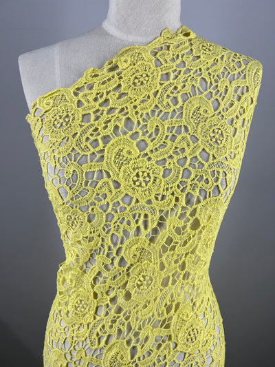 A mannequin is draped with the intricately patterned Anglaise Lace - Limelight from Super Cheap Fabrics. The 100% polyester lace, featuring floral and circular motifs, creates a visually delicate and detailed design. This lightweight lace fabric covers the mannequin asymmetrically, highlighting its texture and craftsmanship.