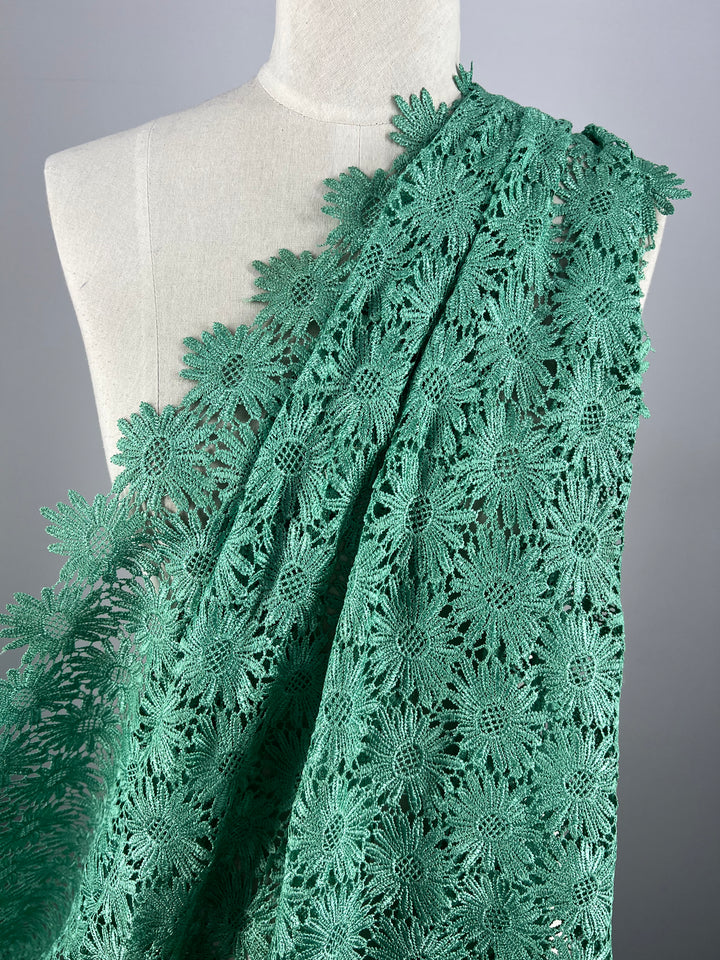 A mannequin is adorned with a large piece of Anglaise Lace in Stone Green from Super Cheap Fabrics, measuring 110cm in width. This exquisite fabric features an intricate floral pattern, perfect for bridal wear. The arrangement highlights the detailed design and delicate craftsmanship against a plain grey background, making the vibrant color and elegant texture of the lace stand out beautifully.