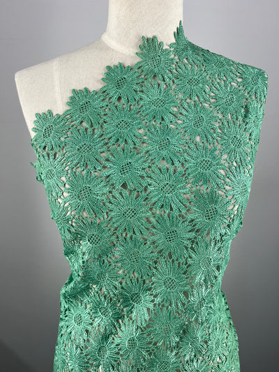 The Anglaise Lace - Stone Green - 110cm draped over a mannequin showcases Super Cheap Fabrics' intricate design. Perfect for bridal wear, the densely arranged floral pattern creates a delicate and elegant texture. The semi-transparent material highlights the exquisite craftwork of this beautiful lace fabric.