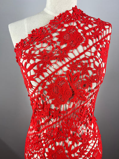 A mannequin is draped in the vibrant Anglaise Lace in Red Alert, a 120cm wide fabric from Super Cheap Fabrics. The intricately crocheted lace with floral patterns wraps around the mannequin in a diagonal fashion, showcasing detailed lacework and open stitches, creating an elegant and delicate appearance reminiscent of high-end bridal wear.