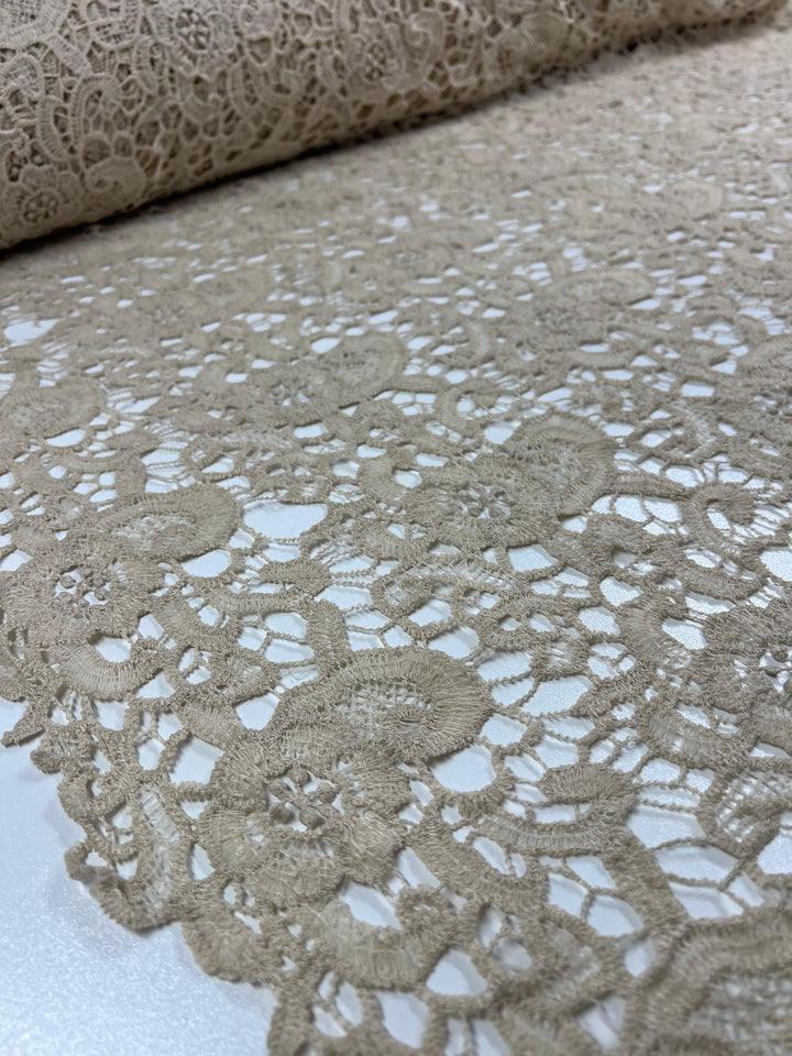 A close-up view of **Anglaise Lace - Wood Ash - 125cm** from **Super Cheap Fabrics**, showcasing its intricate floral patterns. The lace is rolled up in the background and laid out flat in the foreground, highlighting the delicate and detailed texture of the material, perfect for bridal wear or elegant home décor.