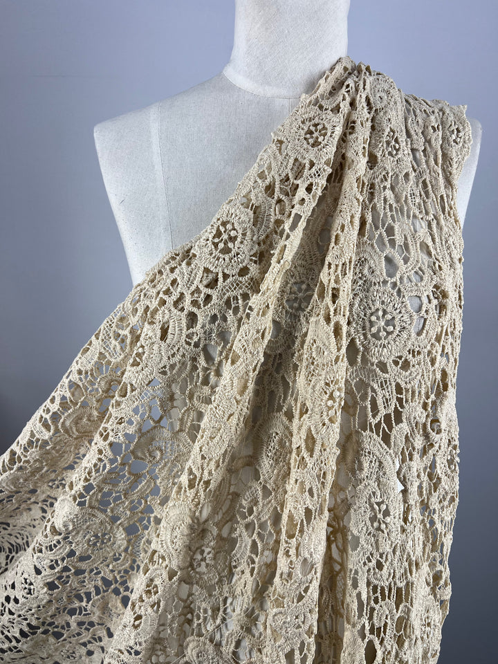 The Anglaise Lace in Wood Ash from Super Cheap Fabrics is draped over a white dress form. This intricately crocheted fabric features various floral and geometric designs, showcasing fine, detailed craftsmanship reminiscent of bridal wear. The background is plain grey, emphasizing the delicate texture of the lace fabrics.