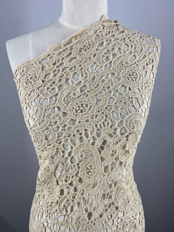 A mannequin is draped with an intricate piece of Super Cheap Fabrics' Anglaise Lace in Wood Ash, measuring 125cm wide. Featuring a detailed floral and geometric pattern, this cream-colored lace is perfect for bridal wear. It is asymmetrical, covering one shoulder and elegantly designed with delicate openwork elements against a plain gray background.