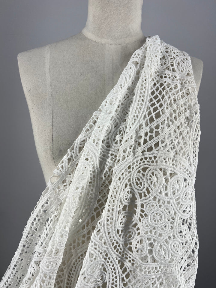 The Anglaise Lace - White - 115cm by Super Cheap Fabrics is gracefully draped over the shoulder of a beige mannequin torso set against a plain gray background. Ideal for bridal wear or home décor, this exquisite white lace boasts intricate and delicate patterns with detailed designs of circles, lines, and floral motifs, creating an elegant and textured appearance.