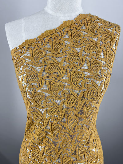 A dress form wearing Super Cheap Fabrics' Anglaise Lace in GOTS Cotton - Yam, a mustard yellow, intricately crocheted garment with a one-shoulder design. Made from lace fabrics, this piece features a detailed leafy floral pattern and semi-transparent sections that create an elegant and delicate appearance. The background is solid light grey.