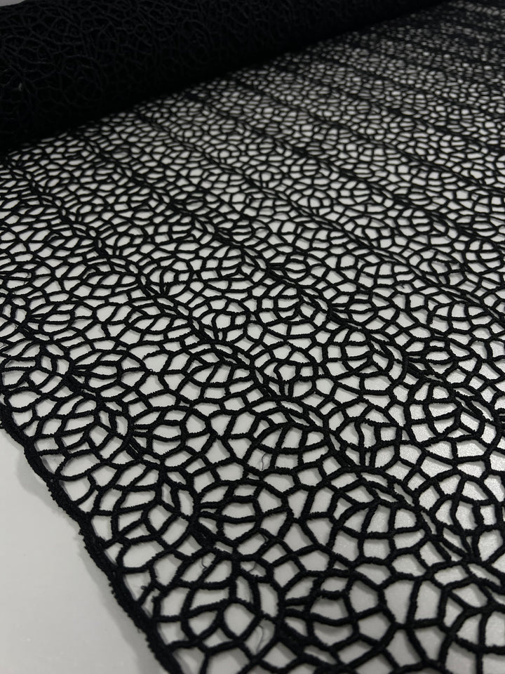 A close-up view of intricately patterned Anglaise Lace - Black - 125cm from Super Cheap Fabrics, rolled out across a surface. The lace fabric features a detailed geometric design with interconnected circular and hexagonal shapes, ideal for bridal wear. The background is a plain, smooth, light-colored surface.