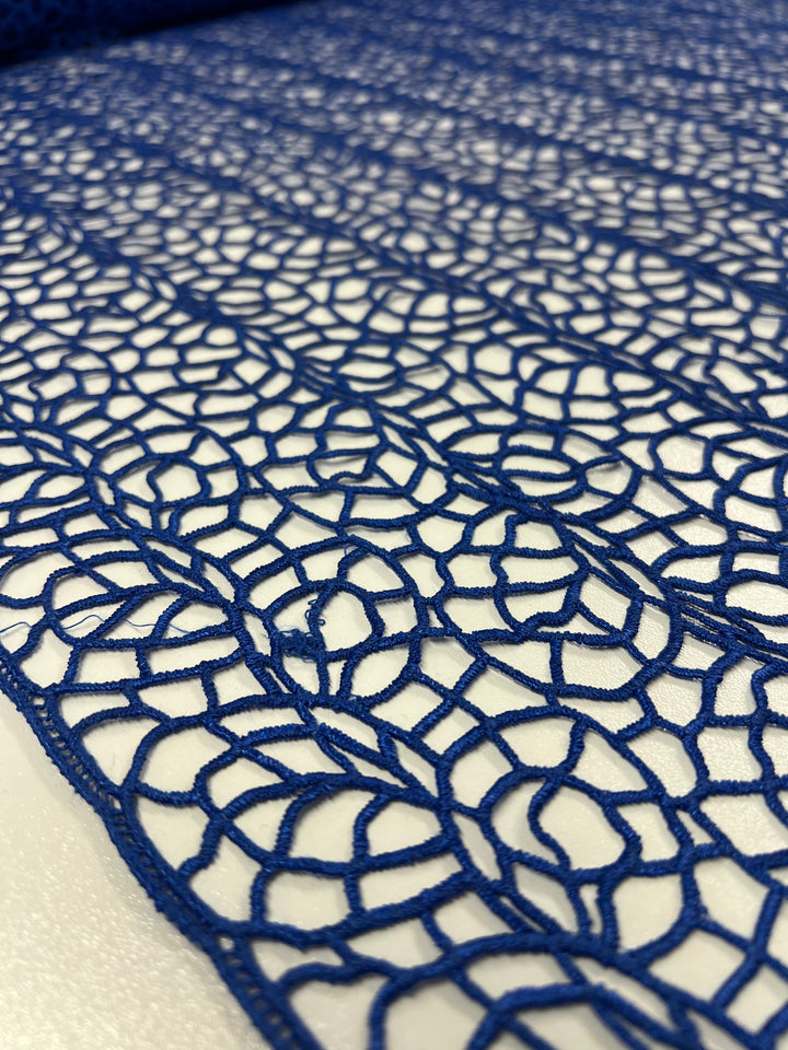 Close-up of Anglaise Lace - Cobalt - 125cm from Super Cheap Fabrics, showcasing intricate circular patterns on a white background. Ideal for home décor or bridal wear, this blue lace fabric weaves together to form an elegant and delicate design, creating a captivating texture. The image highlights the meticulous details and craftsmanship of the fabric.