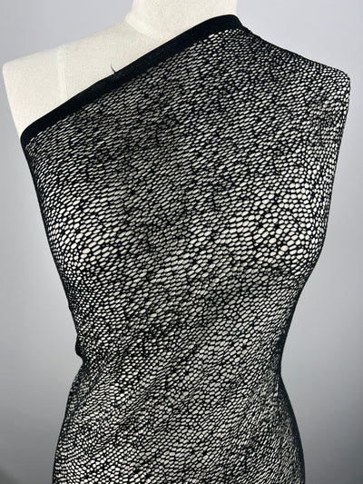 A mannequin is draped in Evening Cobble Stone Lace - Black - 150cm from Super Cheap Fabrics, featuring an intricate, net-like pattern. Made from 100% polyester, this extra lightweight fabric is see-through, revealing the neutral-colored mannequin underneath. The background is plain and grey, keeping the focus on the fabric's texture and design.