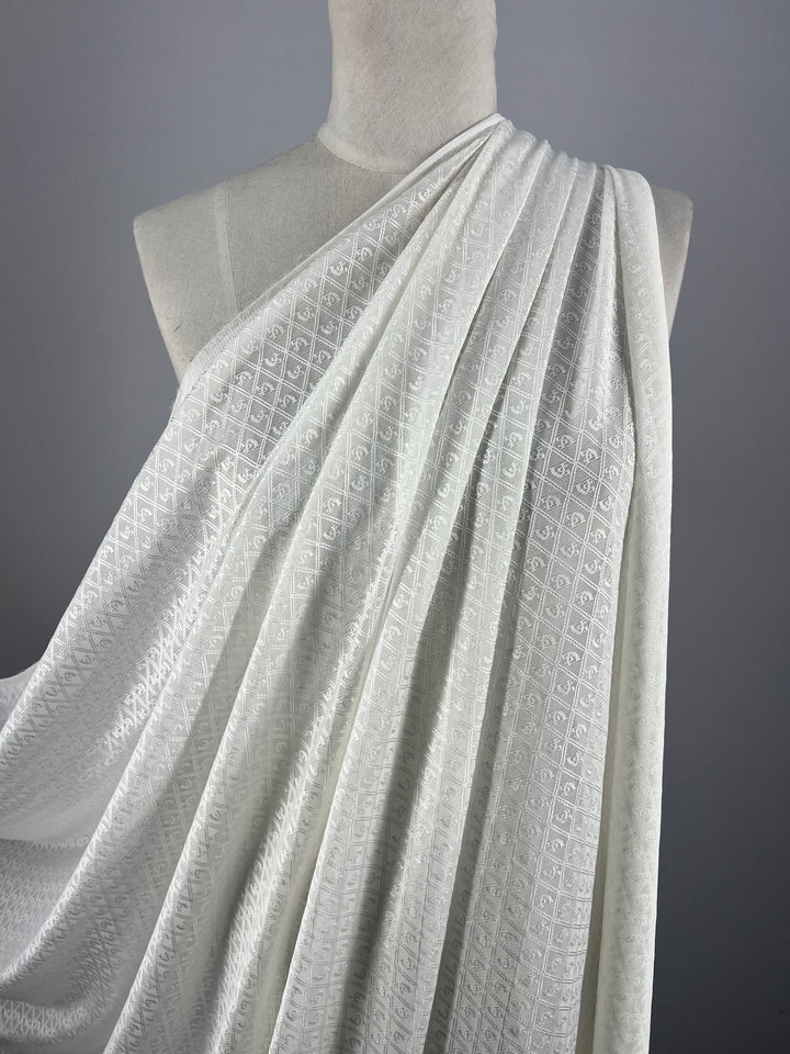 A mannequin draped with Stretch Mesh - Diamond fabric from Super Cheap Fabrics. The 100% nylon, lightweight mesh fabric features a subtle, repetitive design and is arranged gracefully over the shoulder and across the chest of the mannequin. The neutral background highlights the off-white fabric's intricate detail.