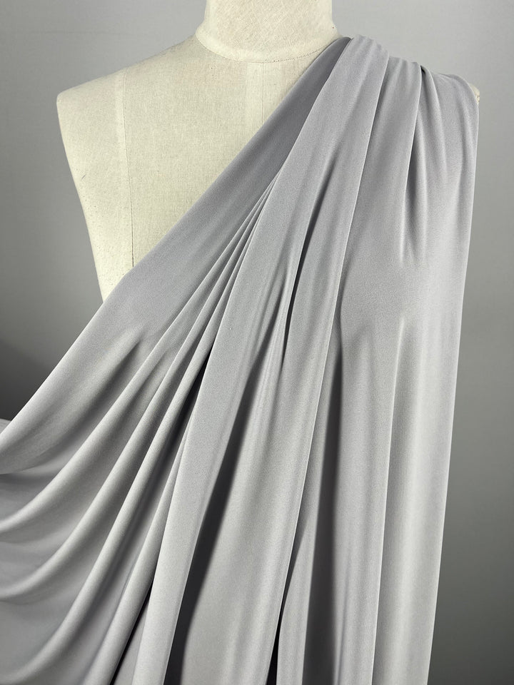 A white mannequin is adorned with Super Cheap Fabrics' ITY Knit in Silver, measuring 150cm. The medium-weight material elegantly flows over one shoulder, showcasing graceful folds and texture. A plain gray background accentuates the simplicity and elegance of the fabric design.