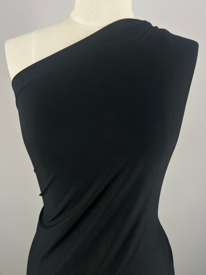 A dress form draped with Super Cheap Fabrics' Cotton Lycra - Black - 160cm, a medium to heavy weight fabric. The material wraps around the form, creating smooth folds and a sleek, elegant appearance. The plain and neutral background ensures that the focus remains on the fabric's texture and draping—ideal for bodycon dresses or high waist skirts.