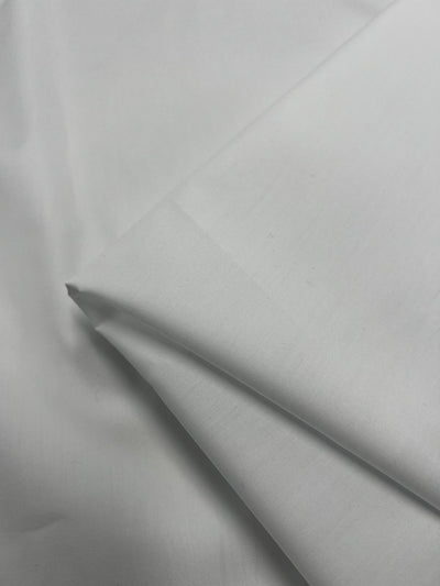 A close-up of the Super Cheap Fabrics' GOTS Cotton - White - 147cm reveals the lightweight material, neatly folded with visible creases. The fabric's smooth texture is emphasized by the lighting, which highlights its gentle folds and edges.