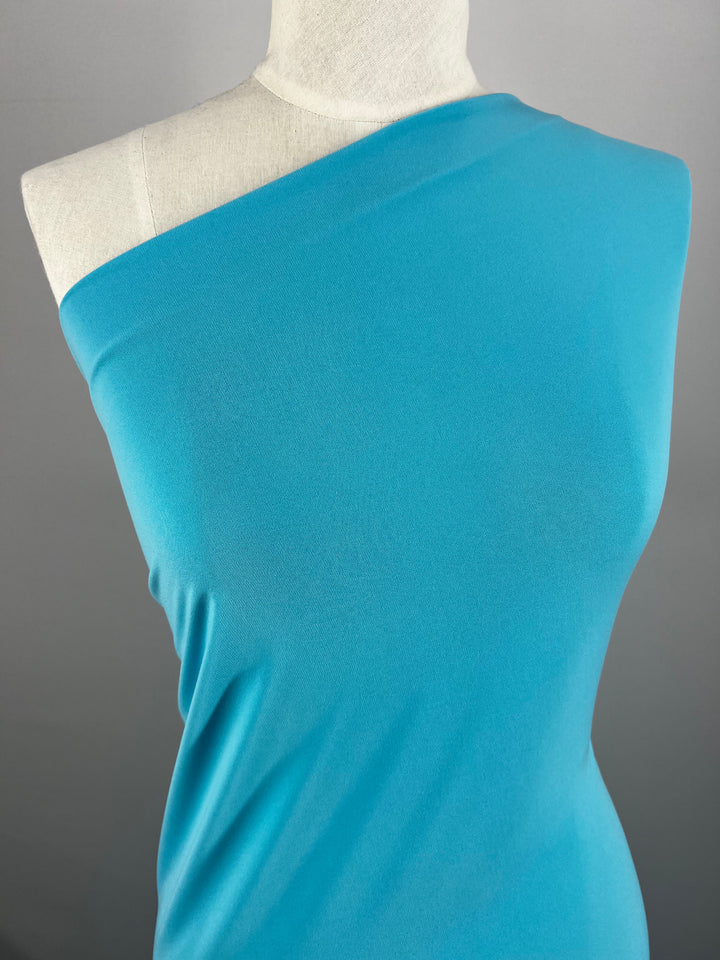 A mannequin presents the "ITY Knit - Capri - 150cm" from Super Cheap Fabrics—a turquoise, one-shoulder dress made from medium-weight fabric—against a plain gray background. The dress drapes elegantly over the mannequin, accentuating its vibrant color and smooth texture.