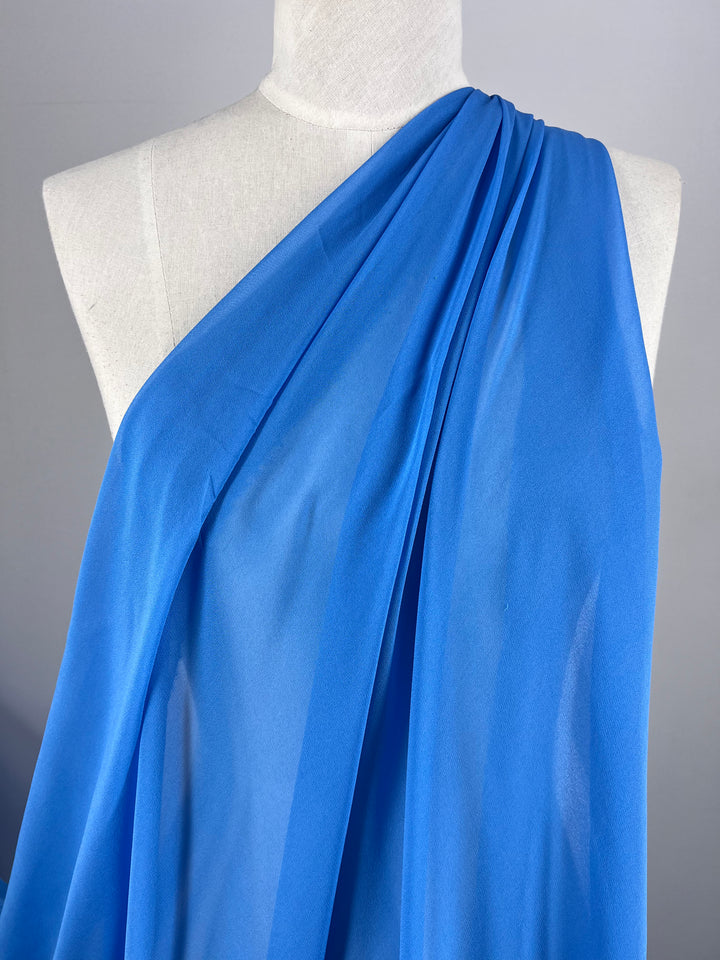 A mannequin draped in Super Cheap Fabrics' Pure Silk Georgette - Denim - 135cm embodies spring-summer style. The vibrant blue one-shoulder fabric is lightweight, gathered at the shoulder, and cascades in soft folds against a plain gray background.