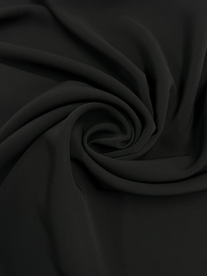 A close-up of Super Cheap Fabrics' Suiting - Saxony Black - 130cm is elegantly draped in a swirl, creating motion and depth. This twill suiting material's texture is smooth and soft, with gentle folds radiating outward from the center.