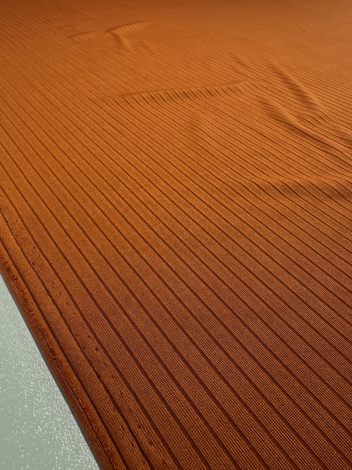 Close-up of the Super Cheap Fabrics 'Rib Knit - Rust - 175cm', a medium weight fabric with parallel rib lines. Its soft, slightly shiny texture features subtle folds and shadows for depth, while its generous stretch enhances flexibility.