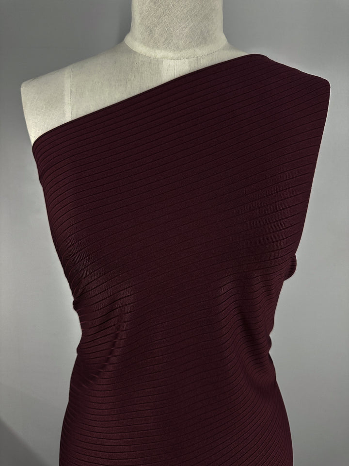 A mannequin displays Super Cheap Fabrics' Rib Knit - Burgundy - 175cm top, featuring textured knit fabrics in a one-shoulder design. The stretch and diagonal rib pattern ensure a snug fit that stands out against a plain gray background.