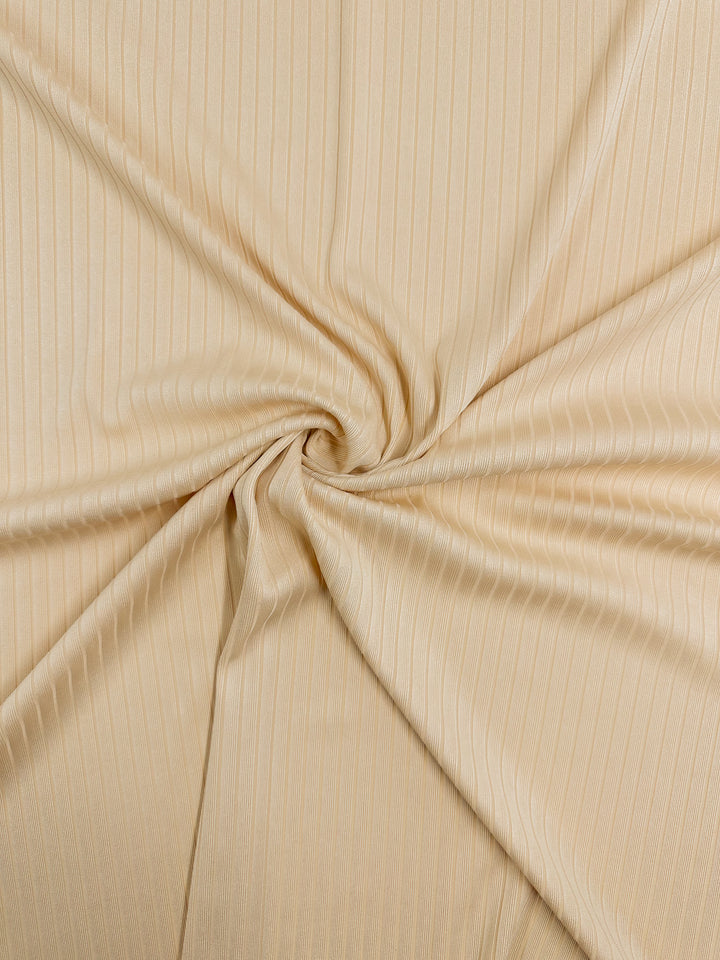 A close-up of Super Cheap Fabrics' Rib Knit - Apricot Gelato - 170cm showcases light beige fabric with subtle vertical pinstripes, swirled into a spiral. This medium weight knit is smooth, slightly glossy, and has gentle folds radiating from the center swirl.