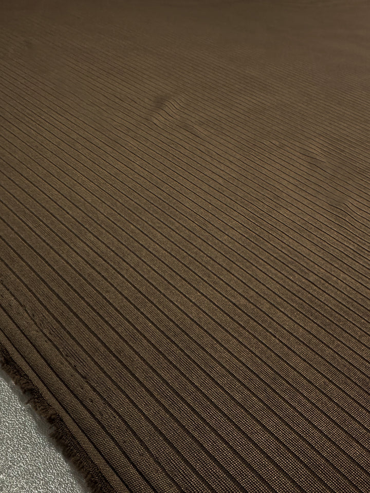 Close-up of "Rib Knit - Pinecone - 170cm" by Super Cheap Fabrics: a dark brown medium weight rib knit with vertical ribbed texture. The smooth material shows evenly spaced lines for a uniform pattern. Visible edges on the left highlight its quality as a textured knit fabric.