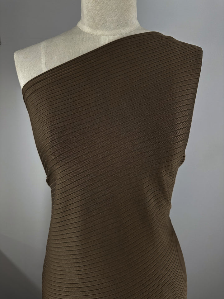 A mannequin displays a ribbed, one-shoulder garment made from Super Cheap Fabrics' medium weight Rib Knit - Pinecone (170cm). This brown fabric is tightly fitted with horizontal ribbing for texture, set against a plain background to highlight the sleek design.