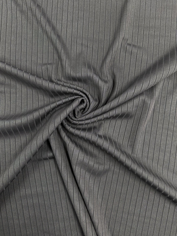 The Rib Knit - Dark Grey - 170cm from Super Cheap Fabrics is shown in a close-up spiral arrangement, displaying its medium weight and subtle sheen that highlights the intricate textured pattern and linear folds.