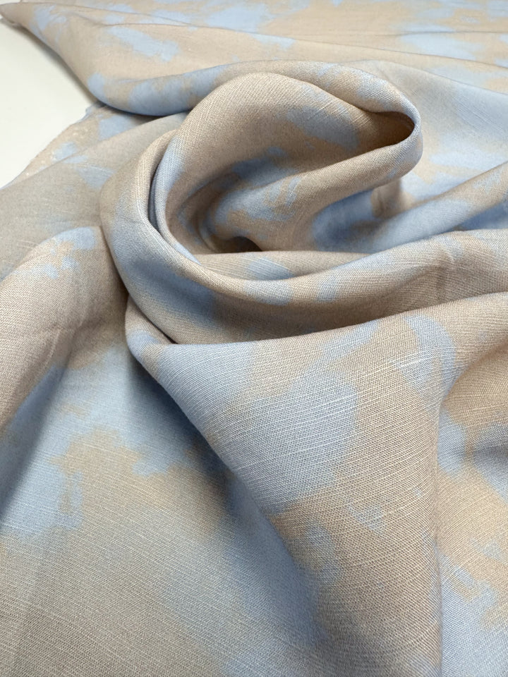 A close-up of the Linen Viscose - Blue Bell - 140cm by Super Cheap Fabrics reveals a soft, flowing fabric with beige and light blue patterns. It drapes naturally for an elegant, serene look, perfect for fancy dresses. Limited stock available.