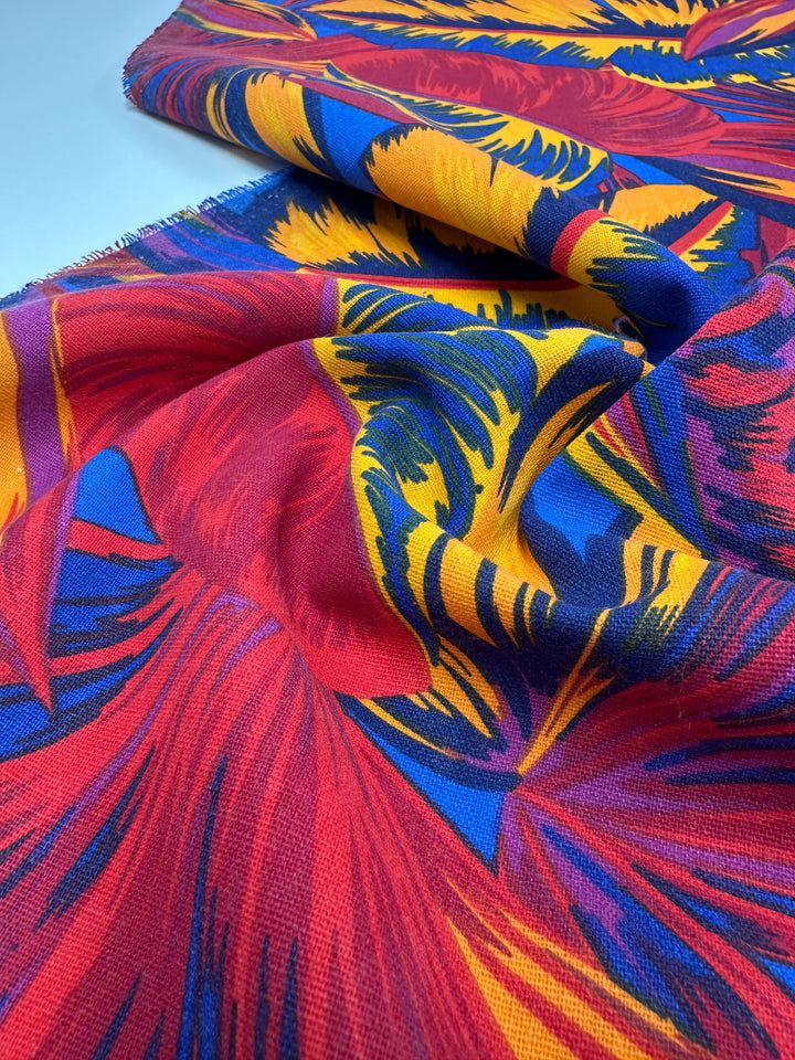 A close-up of Super Cheap Fabrics' Cotton Duck - Palm Scarlet - 140cm features a vivid tropical pattern with red, blue, yellow, and scarlet leaves. Ideal for home décor, the slightly folded fabric creates gentle shadows and highlights.