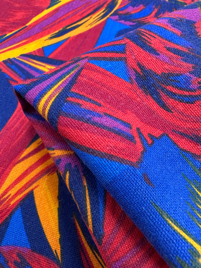 A close-up of Super Cheap Fabrics' Cotton Duck - Palm Scarlet - 140cm fabric reveals its vibrant abstract pattern with bold red, blue, yellow, and flame scarlet strokes. The woven texture makes it ideal for lively home décor.