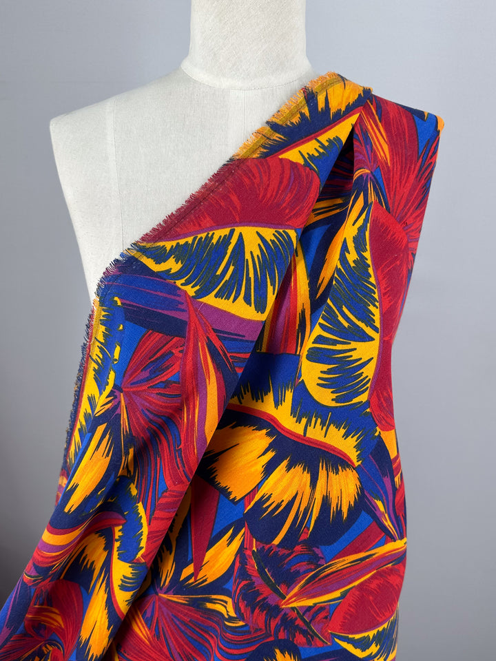 A mannequin displays the vivid Cotton Duck - Palm Scarlet fabric by Super Cheap Fabrics, featuring red, yellow, and Flame Scarlet tropical leaves. The textured cotton with frayed edges stands out against a plain gray backdrop—ideal for inspiring creative home décor.