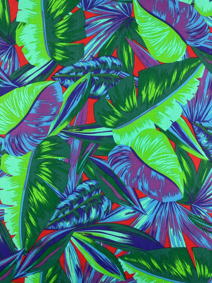 The Cotton Duck - Palm Lush Meadow from Super Cheap Fabrics features a vibrant, jungle-like pattern with overlapping green, blue, and purple tropical leaves on medium-weight cotton against a red backdrop, ideal for lively home décor.