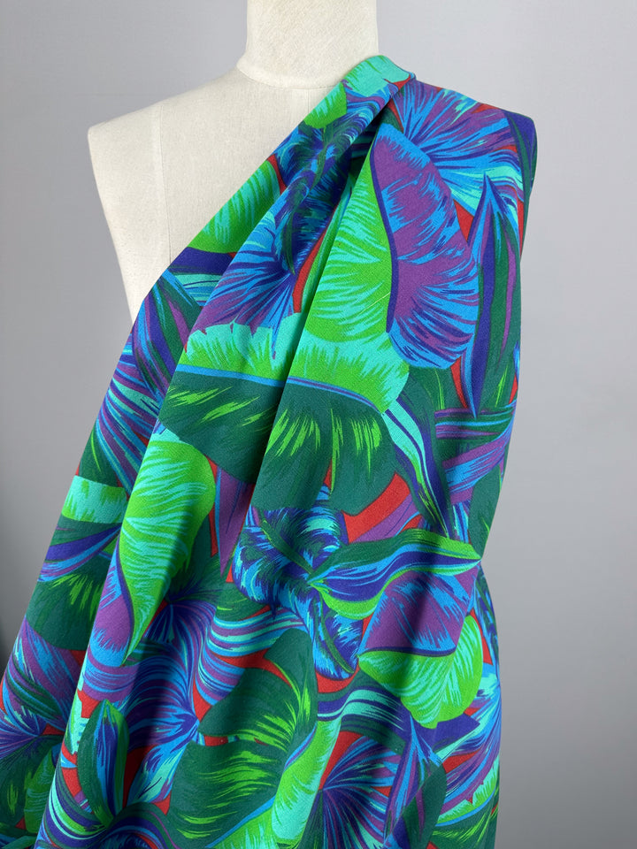 A mannequin displays Super Cheap Fabrics' Cotton Duck in Palm Lush Meadow (140cm), featuring a vibrant tropical leaf pattern in blue, green, and purple on a dark background—ideal for stylish home décor.