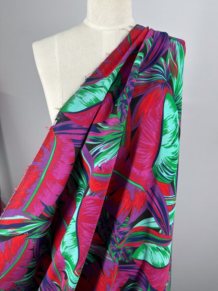 The Super Cheap Fabrics Cotton Duck - Palm Cockatoo, a medium-weight 140cm cotton fabric with vibrant tropical leaf patterns in red, purple, and green, is stylishly draped over a white mannequin, ideal for enhancing home décor.