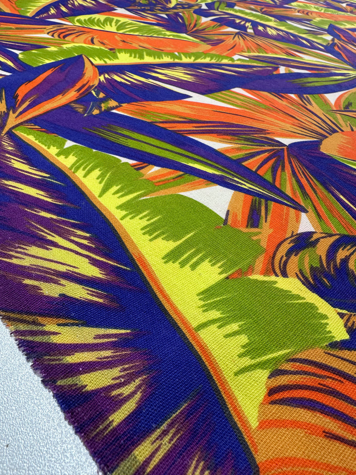 The Cotton Duck - Palm Nectarine by Super Cheap Fabrics is a vibrant cotton fabric featuring tropical leaf patterns in green, orange, purple, and blue. Its dynamic design of overlapping leaves offers a lively touch for home décor. With medium weight, it ensures both durability and versatility.