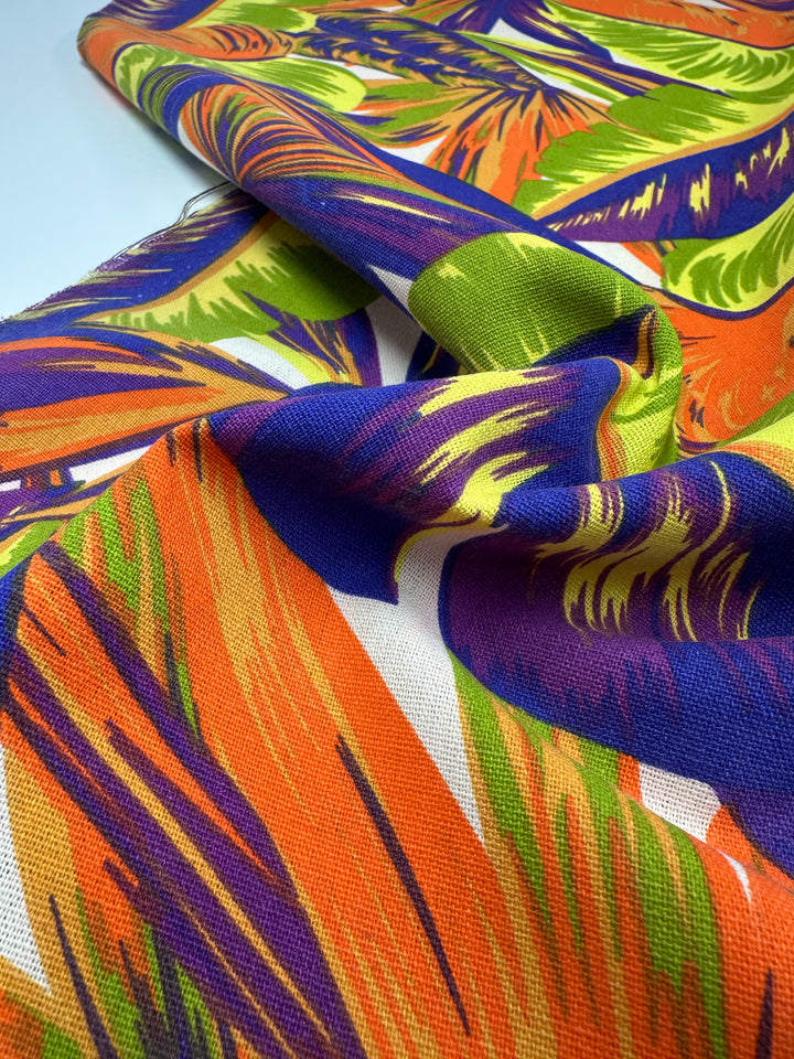 The Cotton Duck - Palm Nectarine by Super Cheap Fabrics is a vibrant tropical cotton fabric featuring a bold pattern of colorful leaves in orange, purple, green, and yellow on a light background. This medium-weight material drapes smoothly with its textured, dynamic form perfect for home décor.