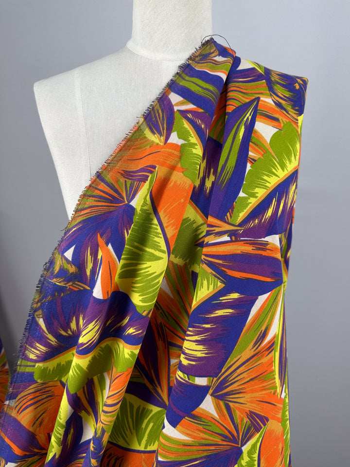 The Cotton Duck - Palm Nectarine fabric by Super Cheap Fabrics is a medium-weight cotton draped on a mannequin, showcasing vibrant tropical leaves in orange, green, purple, and yellow against a neutral backdrop, ideal for bold home décor projects.