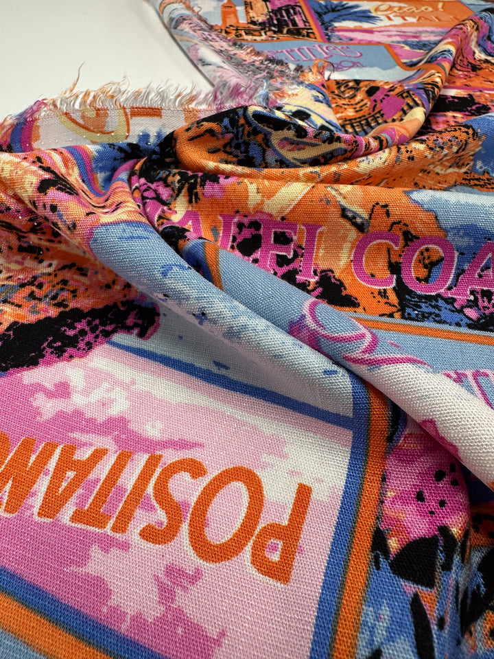 Close-up of a vibrant bamboo rayon fabric from Super Cheap Fabrics with an abstract, dynamic pattern. The "Bamboo Rayon - Italiano - 145cm" features bold text and a mix of orange, blue, pink, and black hues. Frayed edges add texture to this versatile multi-use fabric.