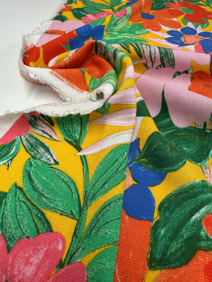 The "Bamboo Rayon - Florita - 145cm" by Super Cheap Fabrics is a lightweight, vibrant multi-use fabric featuring a colorful floral design with red, blue, and green flowers on a yellow and pink background, slightly folded over a plain white surface.