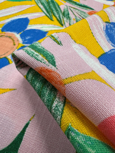 A close-up of Super Cheap Fabrics' Bamboo Rayon - Florita (145cm) highlights a vibrant floral design in vivid blue, yellow, green, pink, and orange hues. This lightweight fabric is folded to showcase its texture and pattern, ideal for versatile projects.