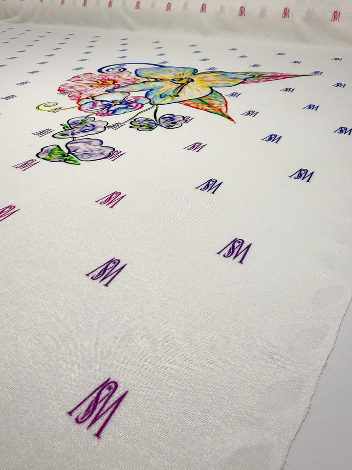 Super Cheap Fabrics' Viscose Knit - Orchid - 135cm is a white viscose knit with a central colorful butterfly and floral print, plus purple “ISM” text embroidered in a grid pattern.