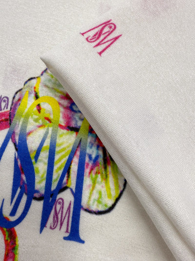 Close-up of Super Cheap Fabrics' Viscose Knit - Orchid (135cm), featuring breathable white fabric with vibrant, artistic prints and overlapping "SM" letters in blue, red, and yellow; fabric is slightly folded to show design details.