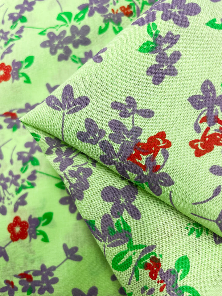 Close-up of Super Cheap Fabrics' Linen Voile - Jazzy - 140cm in light green, featuring a floral pattern. The flowers are primarily purple with some red, accompanied by green leaves. Perfect for home decor, this breathable fabric is shown layered or folded, demonstrating its texture and flexibility.