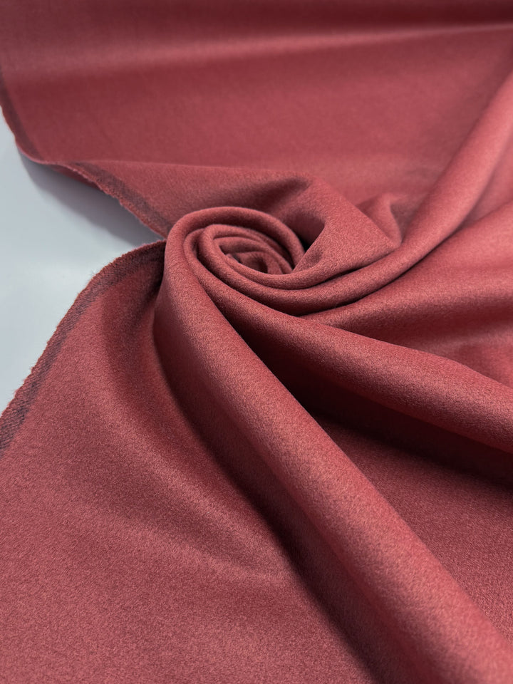 A close-up of Super Cheap Fabrics' Wool Cashmere - Red Ochre - 150cm shows its smooth, velvety texture and gently frayed edges, with the rich red ochre fabric arranged in a soft spiral.